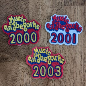 May include: Three embroidered patches with the text "Music in the Parks" and the years 2000, 2001, and 2003. The patches have a red border, blue and yellow text, and are on a wooden surface.