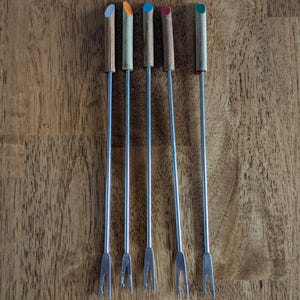 May include: Six fondue forks with wooden handles and colorful tips in blue, orange, green, red, and gray. The metal tines are visible at the bottom of each fork. The forks are arranged on a wooden surface.