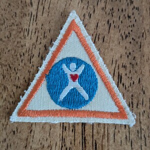 May include: A triangular patch with an orange border and a blue circle in the center. Inside the circle is a white figure with outstretched arms and a red heart. The patch is on a wooden surface.