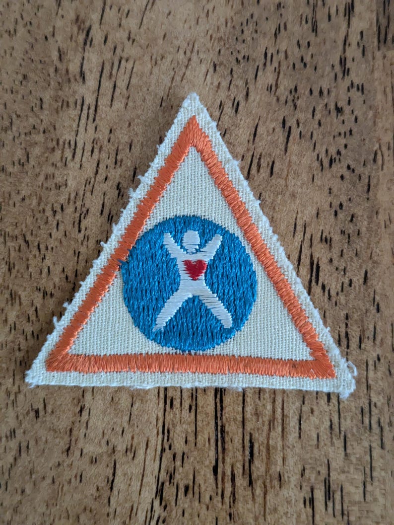 May include: A triangular patch with an orange border and a blue circle in the center. Inside the circle is a white figure with outstretched arms and a red heart. The patch is on a wooden surface.