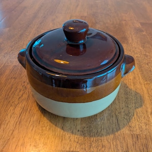 May include: A round ceramic casserole dish with a lid, featuring a two-tone design with a dark brown glazed upper section and a light beige lower section. The pot includes two small side handles and a matching dark brown knob on the lid.