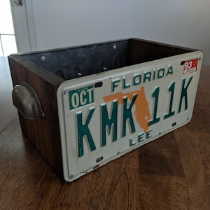 May include: A wooden box with a metal top and a Florida license plate attached to the front. The license plate is white with green lettering and reads "KMK 11K". The license plate also has a small orange sticker that says "Florida" and a red sticker that says "93".