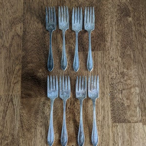 May include: Set of seven silver-plated dessert forks with ornate designs. The forks are arranged in two rows on a wooden surface.