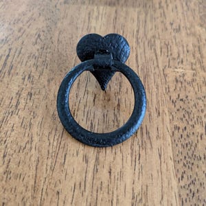May include: Black metal drawer pull with a heart-shaped backplate and a circular handle. The hardware has a textured surface and is set against a wooden background. This decorative pull is suitable for cabinets and furniture.