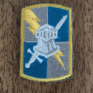 May include: Embroidered patch with a shield design. The patch features a white helmet, sword, and lightning bolts on a blue and gray background, outlined in yellow. The patch is rectangular with rounded corners.