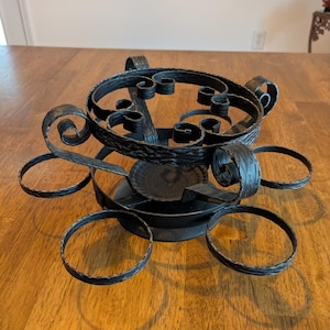 May include: A black metal stand featuring a textured, hammered finish with a central circular base and six radiating ring-shaped holders. The structure includes decorative scrollwork elements connecting the upper ring to the base, creating a multi-tiered appearance for holding small items.