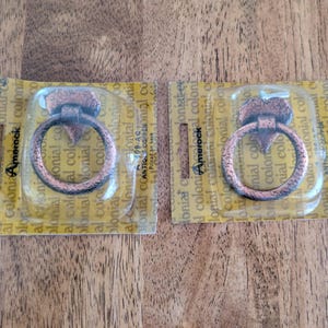 May include: Two antique copper-colored metal ring pulls, each mounted on a rectangular cardboard backing with clear plastic packaging. The pulls feature a circular ring attached to a heart-shaped base, branded with the Amerock Colonial logo.