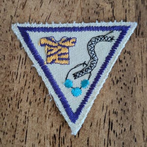 May include: Triangular patch with a cream-colored background, a purple border, and a purple outline. The patch features a yellow and purple shirt, a black and white snake, and three turquoise beads. The patch is likely for a youth organization.