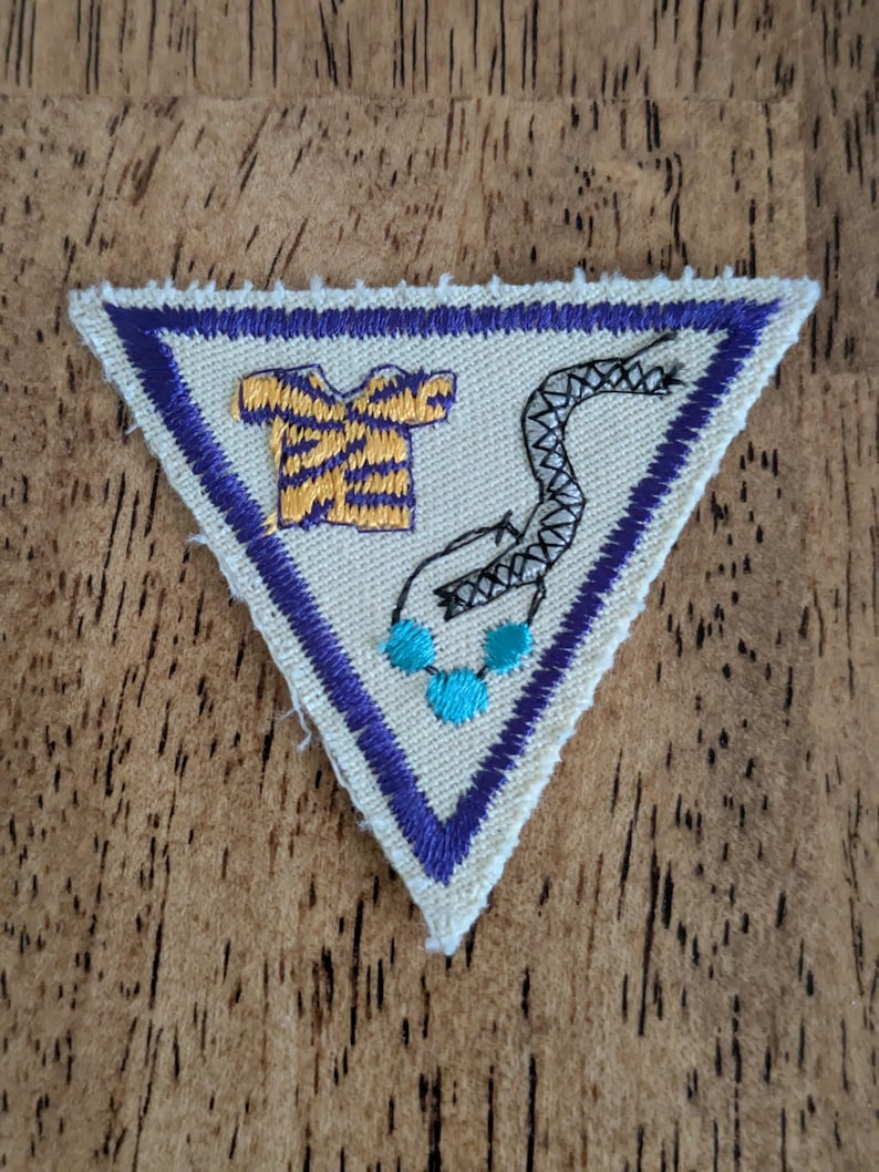 May include: Triangular patch with a cream-colored background, a purple border, and a purple outline. The patch features a yellow and purple shirt, a black and white snake, and three turquoise beads. The patch is likely for a youth organization.
