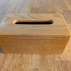 May include: A rectangular tissue box cover crafted from light brown wood with a visible grain pattern. The cover features a smooth, tapered design with a centered, elongated oval opening on the top surface for dispensing tissues.