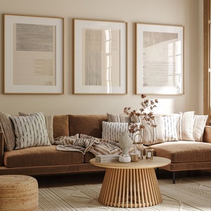 Könnte beinhalten: Ein braunes Ecksofa mit einem Holz-Couchtisch und einem beigefarbenen Teppich. Das Sofa hat mehrere Kissen und eine Decke. An der Wand hinter dem Sofa hängen drei gerahmte abstrakte Kunstwerke.