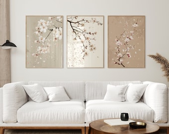 May include: Three framed prints of delicate white cherry blossoms with pink accents on a beige background. The prints are hanging on a white wall above a white sofa with pillows.