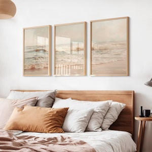 May include: Three framed prints of ocean waves crashing on a sandy beach. The prints are in a light brown frame and are hung on a white wall above a bed with a brown headboard.