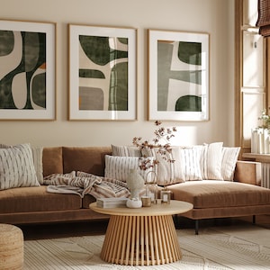 Op de afbeelding: Een bruine hoekbank met een deken en kussens in een woonkamer met drie ingelijste abstracte kunstprints met groene en bruine geometrische vormen. De bank staat voor een raam met een witte vensterbank en een witte radiator.