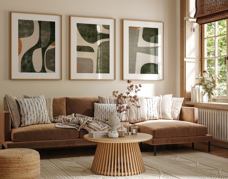 Op de afbeelding: Een bruine hoekbank met een deken en kussens in een woonkamer met drie ingelijste abstracte kunstprints met groene en bruine geometrische vormen. De bank staat voor een raam met een witte vensterbank en een witte radiator.
