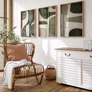 Op de afbeelding: Een lichtbruine rieten stoel met een wit deken en een bruin kussen. Drie ingelijste abstracte kunstprints in groen, wit en bruin hangen aan de muur achter de stoel. Een witte kast met een houten blad en twee lades bevindt zich op de achtergrond.