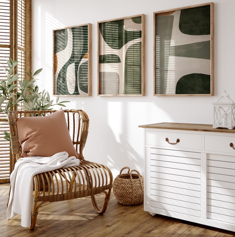 Op de afbeelding: Een lichtbruine rieten stoel met een wit deken en een bruin kussen. Drie ingelijste abstracte kunstprints in groen, wit en bruin hangen aan de muur achter de stoel. Een witte kast met een houten blad en twee lades bevindt zich op de achtergrond.