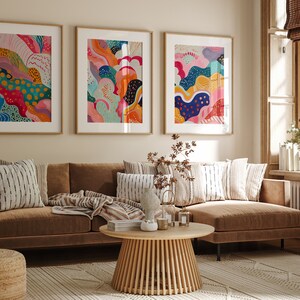 May include: Three framed abstract art prints with colorful, swirling patterns in shades of pink, blue, yellow, orange, and green. The prints are hanging on a beige wall above a brown sectional sofa with striped throw pillows.