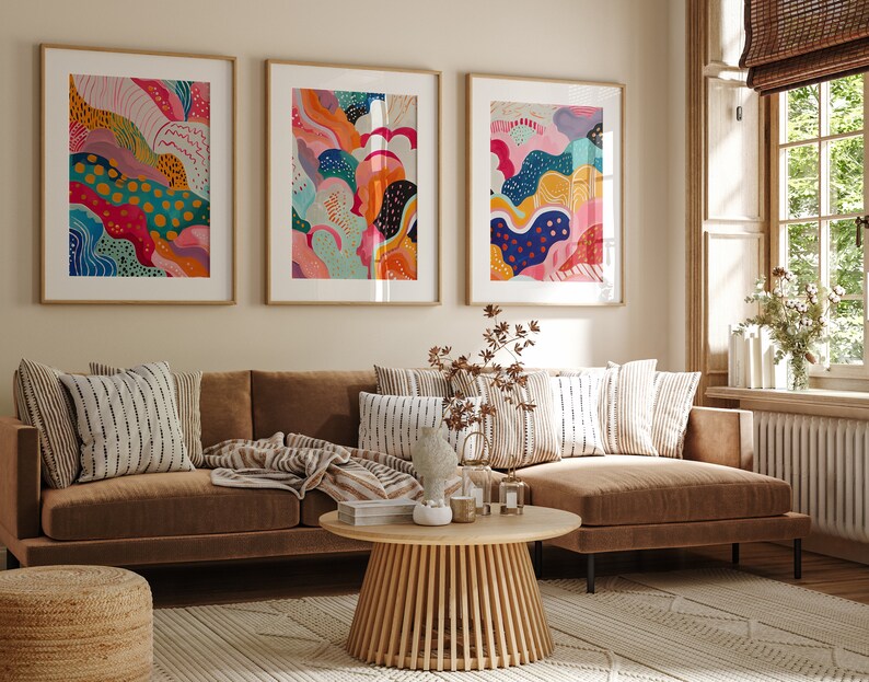 May include: Three framed abstract art prints with colorful, swirling patterns in shades of pink, blue, yellow, orange, and green. The prints are hanging on a beige wall above a brown sectional sofa with striped throw pillows.