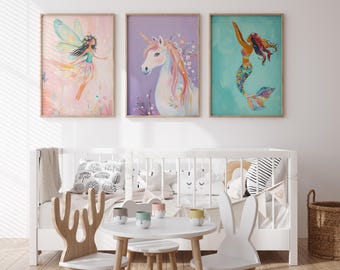 May include: Three framed illustrations of a fairy, a unicorn, and a mermaid. The fairy is wearing a pink dress and has green wings. The unicorn is white with a pink mane and tail. The mermaid is wearing a blue and green tail and has brown hair. The illustrations are hanging on a white wall above a white crib with a white table and chairs in front of it.