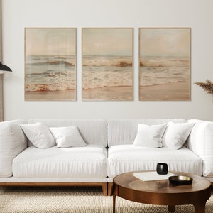 May include: Three framed prints of ocean waves crashing on a sandy beach. The prints are in a light brown frame and have a vintage, faded look. The prints are hanging on a white wall above a white couch with pillows.