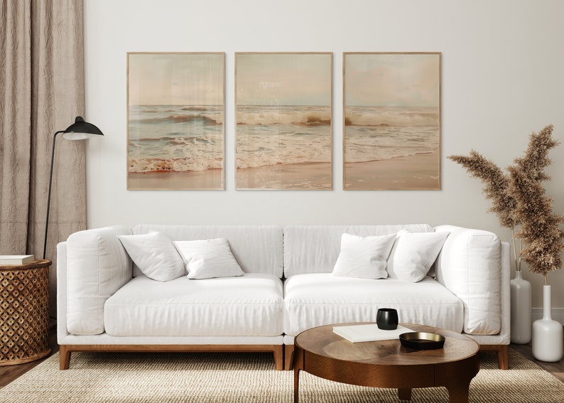 May include: Three framed prints of ocean waves crashing on a sandy beach. The prints are in a light brown frame and have a vintage, faded look. The prints are hanging on a white wall above a white couch with pillows.