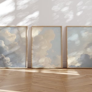 May include: Three framed prints of a cloudy sky with a light blue and beige color scheme. The prints are arranged in a row on a wooden floor.