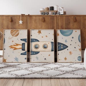 May include: Three framed art prints with a space theme. Each print features a rocket ship, planets, and stars in shades of blue, orange, and beige. The prints are displayed on a light-coloured rug in front of a wooden cabinet.