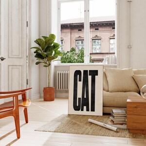 May include: A framed art print with the word "CAT" in bold, black letters. The print is displayed in a bright, airy living room setting, alongside a beige sofa, wooden furniture, and a potted plant. A rug and a window are also visible.