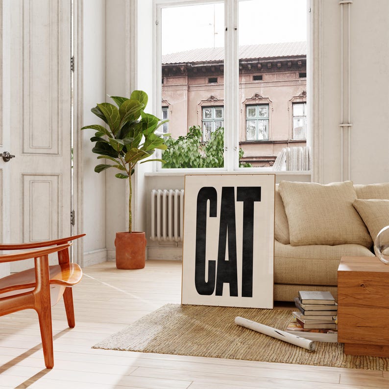 May include: A framed art print with the word "CAT" in bold, black letters. The print is displayed in a bright, airy living room setting, alongside a beige sofa, wooden furniture, and a potted plant. A rug and a window are also visible.