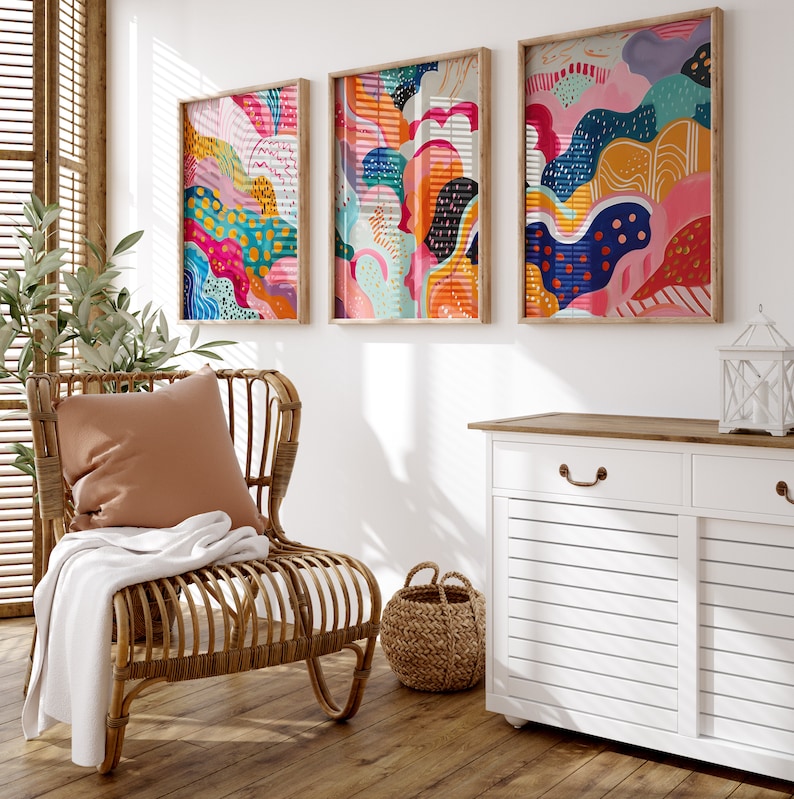 May include: Three framed abstract paintings with colorful, organic shapes and patterns in shades of pink, blue, yellow, and orange. The paintings are hanging on a white wall above a white cabinet with two drawers.