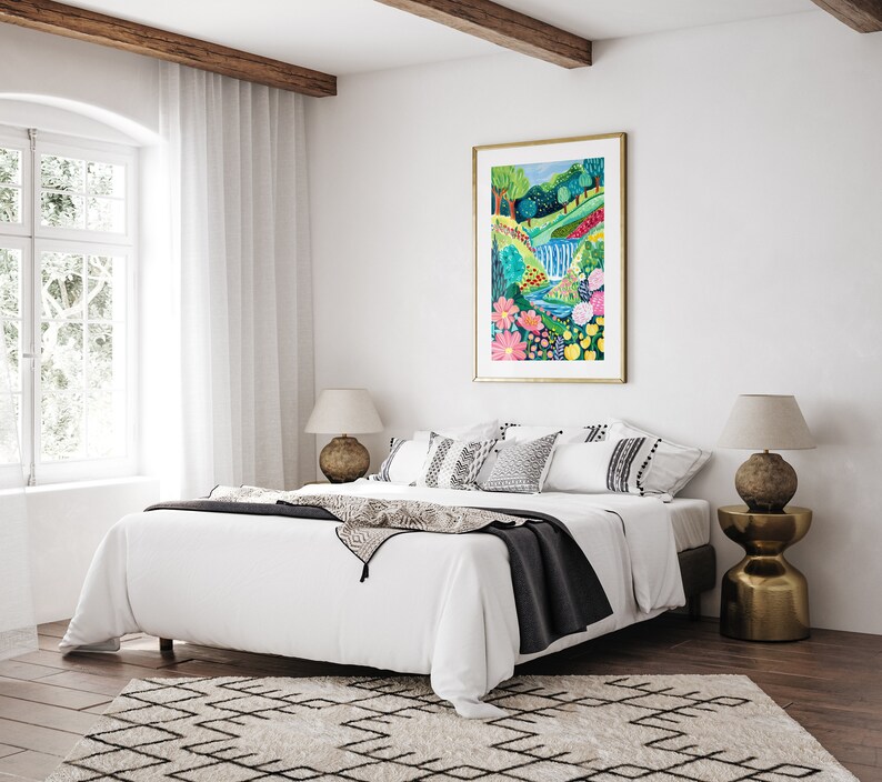 May include: A white bed with a black throw blanket and patterned pillows in a bedroom with a large painting of a colorful landscape with a waterfall and flowers above the bed. Two lamps with gold bases are on nightstands on either side of the bed.