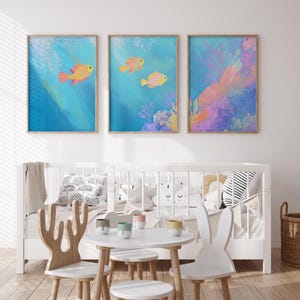 May include: Three framed illustrations of a coral reef scene with yellow and pink fish swimming in blue water. The illustrations are hung on a white wall above a white crib with a white and black striped pillow and a white blanket.