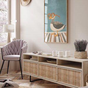 May include: A framed art print featuring a cartoon goose wearing orange glasses. The artwork hangs above a light wood cabinet with woven rattan doors. A lavender plant in a woven basket sits on the cabinet. A light purple chair and a hat are also visible.