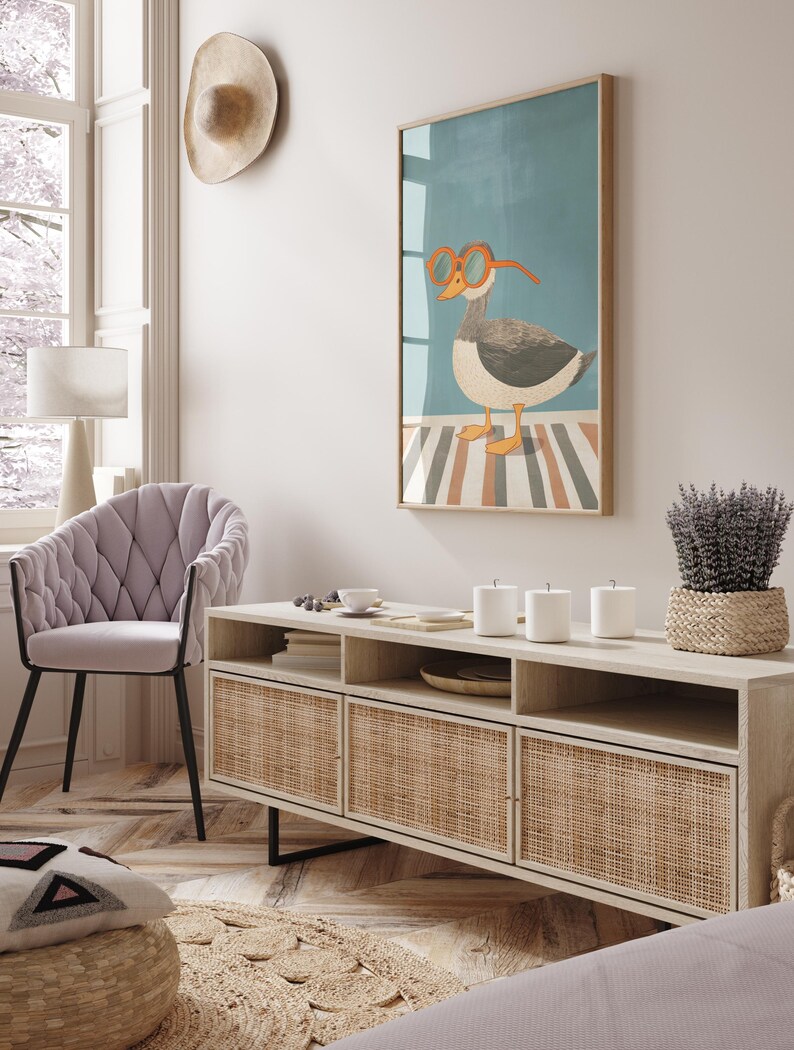 May include: A framed art print featuring a cartoon goose wearing orange glasses. The artwork hangs above a light wood cabinet with woven rattan doors. A lavender plant in a woven basket sits on the cabinet. A light purple chair and a hat are also visible.