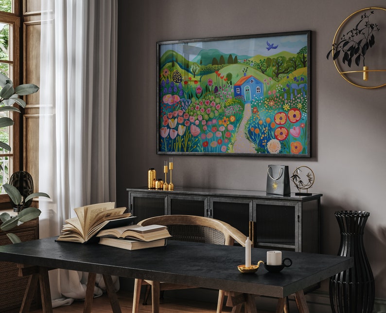 May include: A colorful painting of a blue house in a field of flowers. The house has a white door and a red roof. The flowers are in shades of pink, red, yellow, and orange. The painting is framed in black and hangs on a gray wall.
