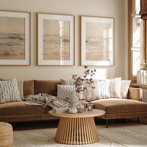 May include: A brown sectional sofa with a throw blanket and pillows in a living room with three framed pictures of seascapes above the sofa. The sofa is in front of a window with a view of trees and a white radiator.