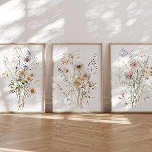 以下が含まれることがあります： 花束を描いた水彩画が3枚、額装されています。それぞれの絵は、白、ピンク、青、黄色の花々を異なる配置で描いています。