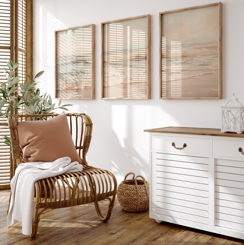 May include: A light brown wicker chair with a tan throw blanket and a peach-colored pillow sits in a room with three framed beach-themed prints and a white cabinet with a wood top.