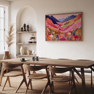 K&ouml;nnte beinhalten: Ein gerahmtes abstraktes Landschaftsbild mit einer farbenfrohen Bergkette und Wildblumen in Rosa-, Orange-, Gelb- und Blaut&ouml;nen. Das Bild h&auml;ngt an einer wei&szlig;en Wand &uuml;ber einem Esstisch aus Holz mit St&uuml;hlen.