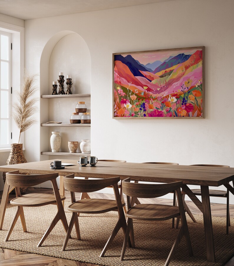 K&ouml;nnte beinhalten: Ein gerahmtes abstraktes Landschaftsbild mit einer farbenfrohen Bergkette und Wildblumen in Rosa-, Orange-, Gelb- und Blaut&ouml;nen. Das Bild h&auml;ngt an einer wei&szlig;en Wand &uuml;ber einem Esstisch aus Holz mit St&uuml;hlen.