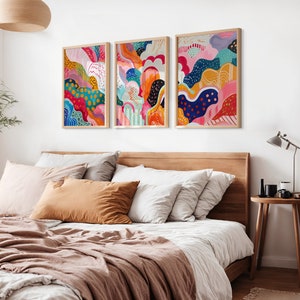 May include: Three abstract art prints in wooden frames, each featuring a colorful, swirling design with a mix of pink, blue, green, yellow, orange, and black. The prints are hung on a white wall above a wooden bed frame.
