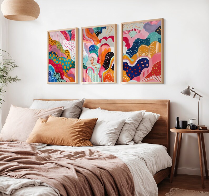 May include: Three abstract art prints in wooden frames, each featuring a colorful, swirling design with a mix of pink, blue, green, yellow, orange, and black. The prints are hung on a white wall above a wooden bed frame.