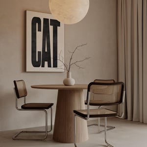 May include: A modern dining room setup featuring a round wooden table and four chairs with woven backs and dark seats. A framed artwork with the word "CAT" in bold black letters hangs on the wall. A white vase with branches sits on the table.
