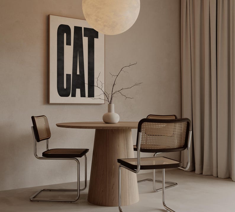 May include: A modern dining room setup featuring a round wooden table and four chairs with woven backs and dark seats. A framed artwork with the word "CAT" in bold black letters hangs on the wall. A white vase with branches sits on the table.