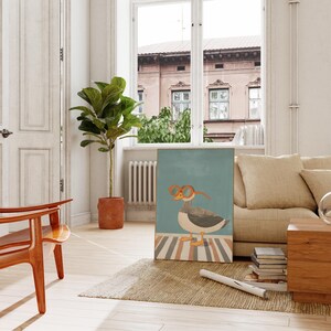 May include: A framed art print featuring a whimsical illustration of a goose wearing orange glasses. The goose stands on a striped surface against a teal background. The artwork is displayed in a bright, airy room with a sofa and a potted plant.
