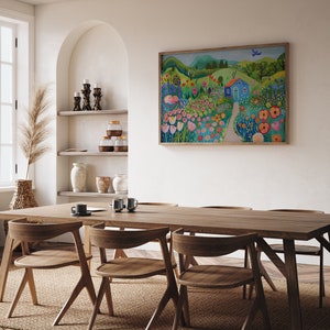 May include: A painting of a colorful landscape with a blue house, flowers, and a bird in the sky. The painting is framed and hanging on a white wall in a dining room with a wooden table and chairs.