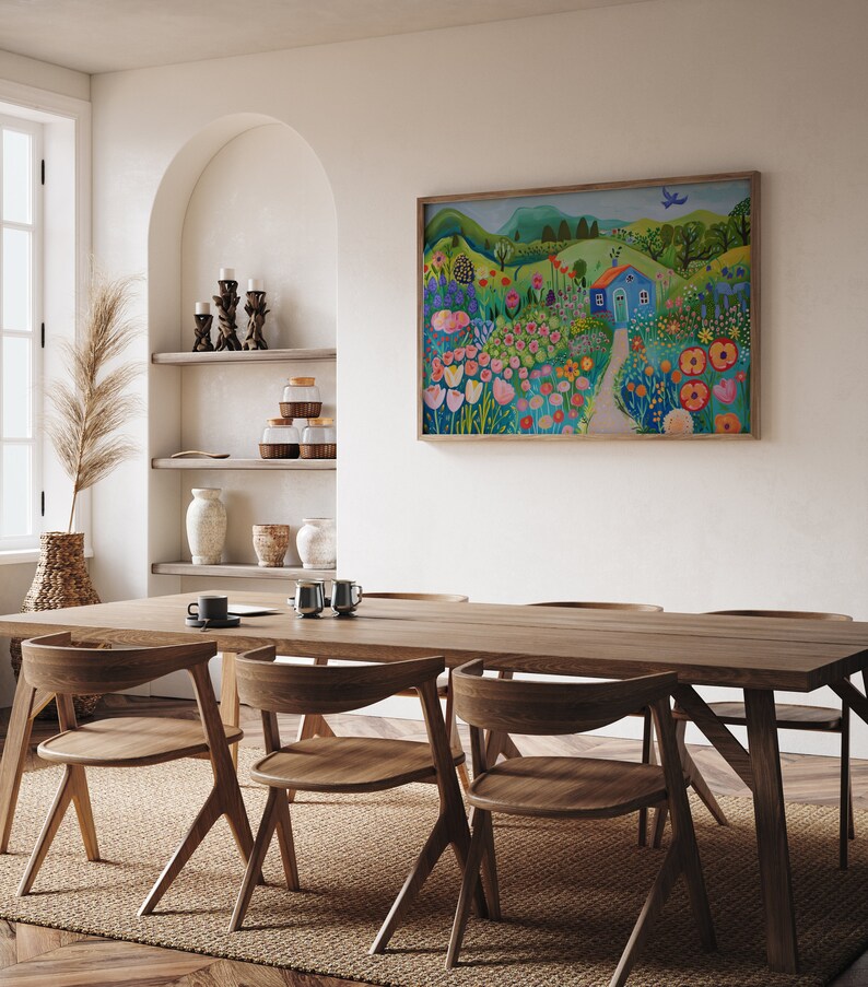 May include: A painting of a colorful landscape with a blue house, flowers, and a bird in the sky. The painting is framed and hanging on a white wall in a dining room with a wooden table and chairs.