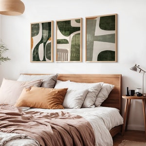 Op de afbeelding: Drie abstracte kunstprints in een slaapkameromgeving. De prints hebben een groen-wit kleurenschema met geometrische vormen. De prints zijn ingelijst in een licht houten lijst en hangen aan een witte muur boven een bed met een bruin hoofdeinde.