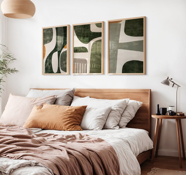 Op de afbeelding: Drie abstracte kunstprints in een slaapkameromgeving. De prints hebben een groen-wit kleurenschema met geometrische vormen. De prints zijn ingelijst in een licht houten lijst en hangen aan een witte muur boven een bed met een bruin hoofdeinde.